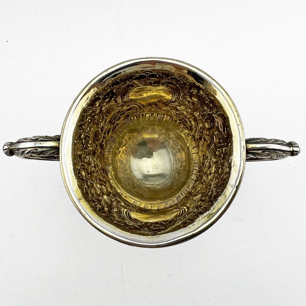 A George III sterling silver campana-shaped cup featuring chased floral motifs, a vacant cartouche, twin scroll handles, and a gilt interior. View IV