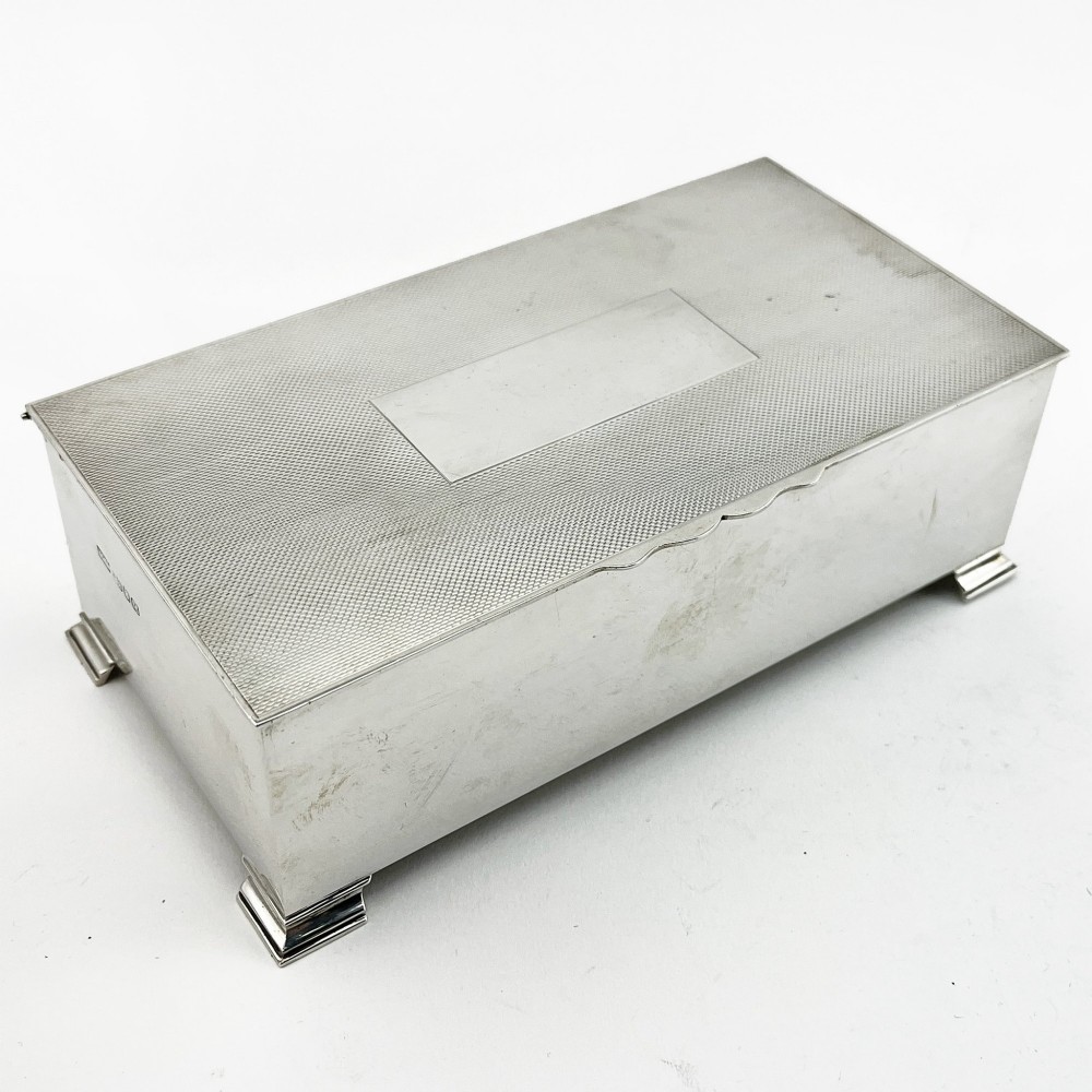 A rectangular sterling silver box featuring an engine-turned patterned lid, a central rectangular cartouche, and four stepped bracket feet.
