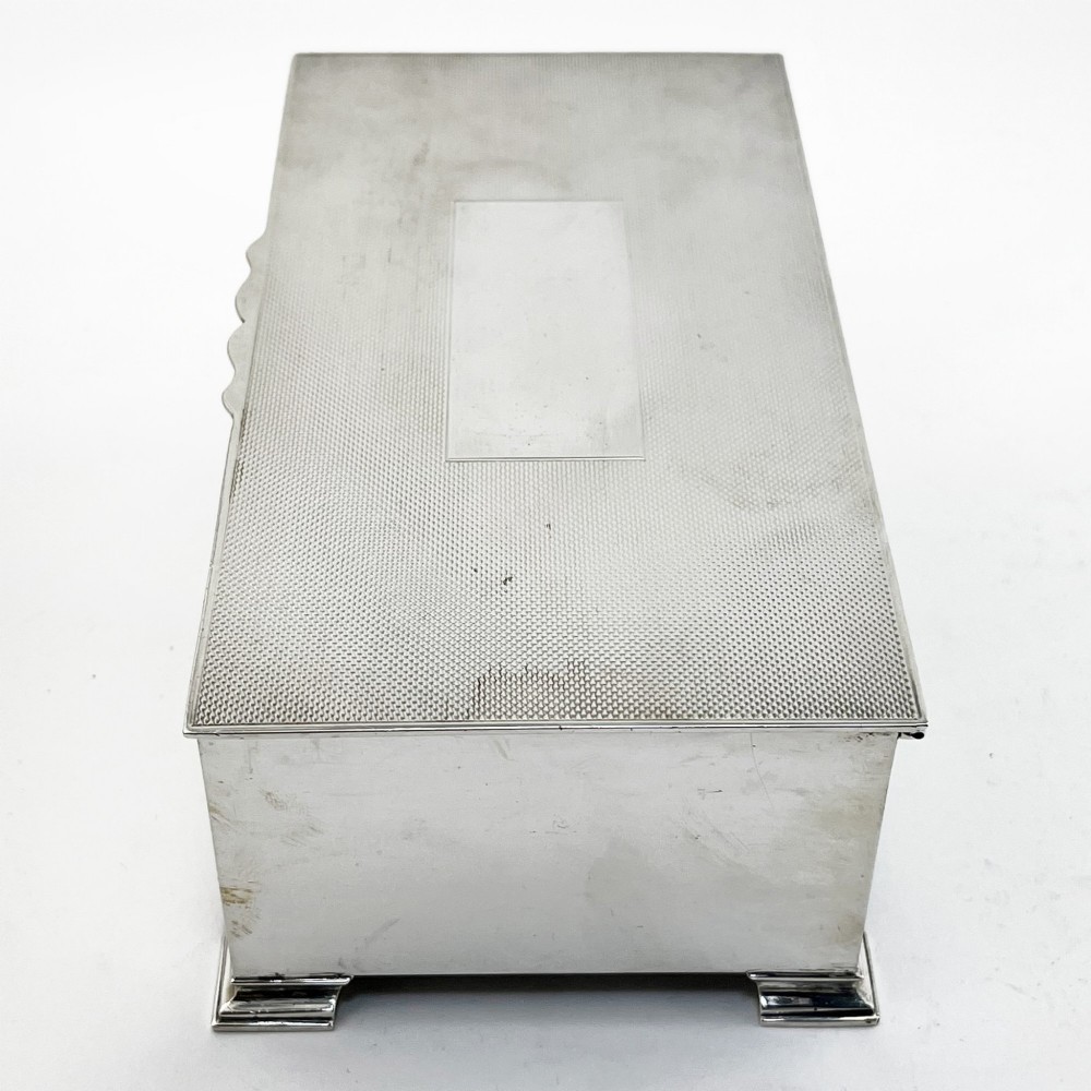 A rectangular sterling silver box featuring an engine-turned patterned lid, a central rectangular cartouche, and four stepped bracket feet. View II