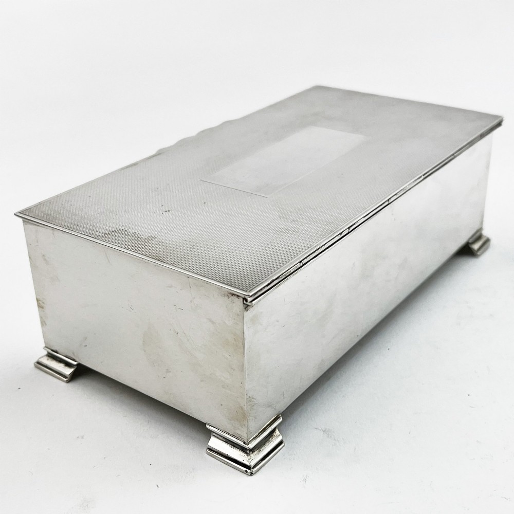 A rectangular sterling silver box featuring an engine-turned patterned lid, a central rectangular cartouche, and four stepped bracket feet. View III
