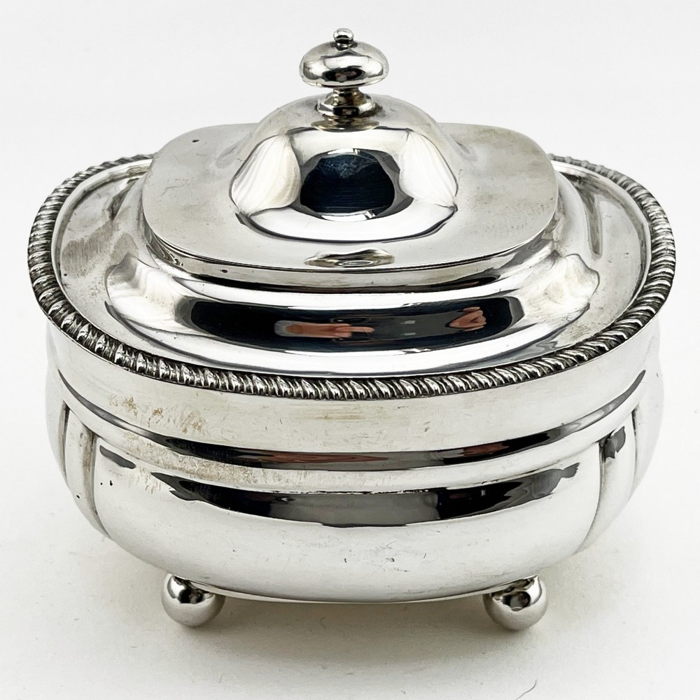 A polished sterling silver tea caddy featuring a lobed body, ropework border trim, domed lid with finial, and four small ball feet.