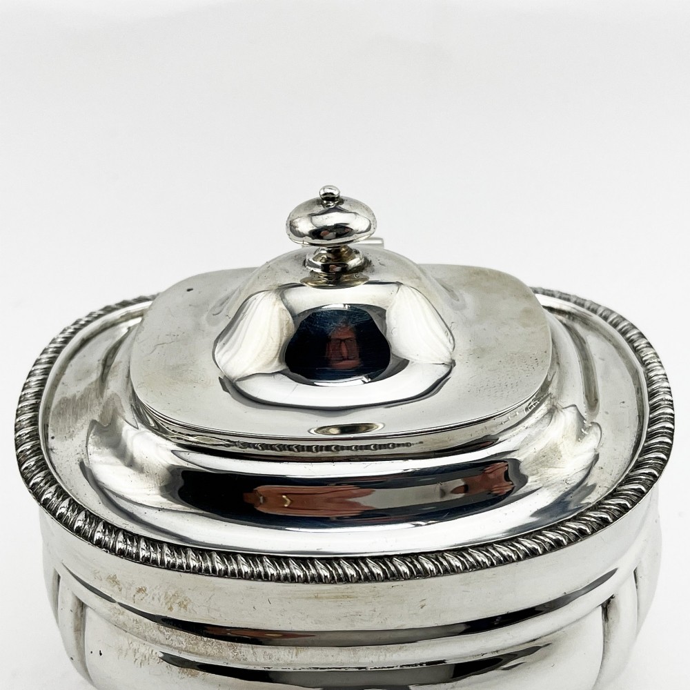 A polished sterling silver tea caddy featuring a lobed body, ropework border trim, domed lid with finial, and four small ball feet. View II