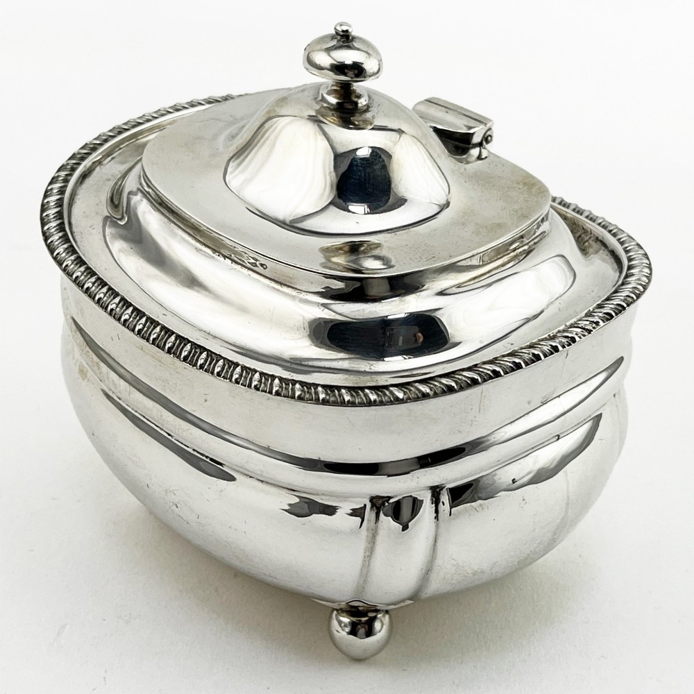 A polished sterling silver tea caddy featuring a lobed body, ropework border trim, domed lid with finial, and four small ball feet. View III