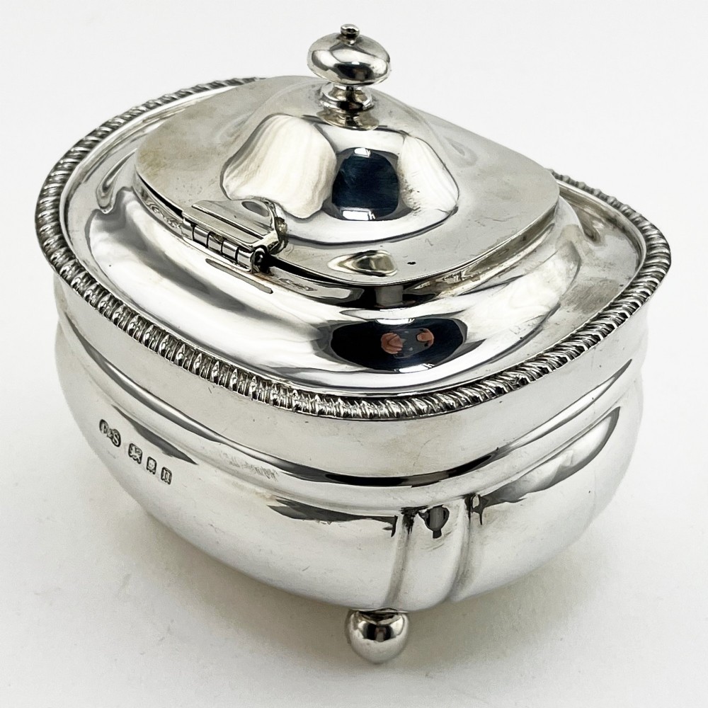 A polished sterling silver tea caddy featuring a lobed body, ropework border trim, domed lid with finial, and four small ball feet. View IV