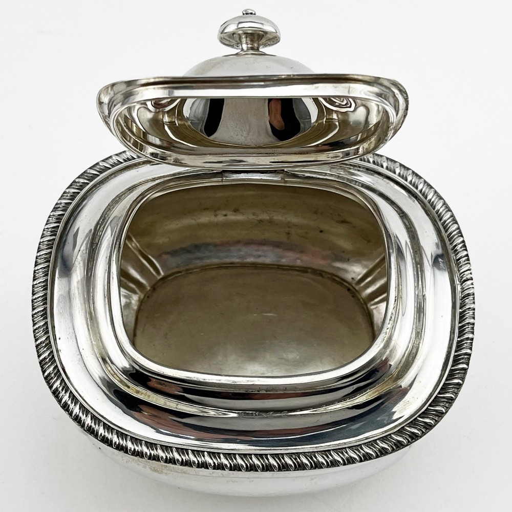 A polished sterling silver tea caddy featuring a lobed body, ropework border trim, domed lid with finial, and four small ball feet. View V