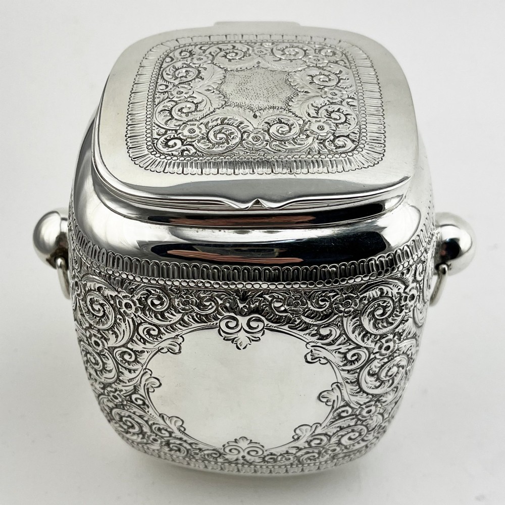 A silver-plated rectangular tea caddy featuring elaborate floral engraving, side handles, and a central blank cartouche.