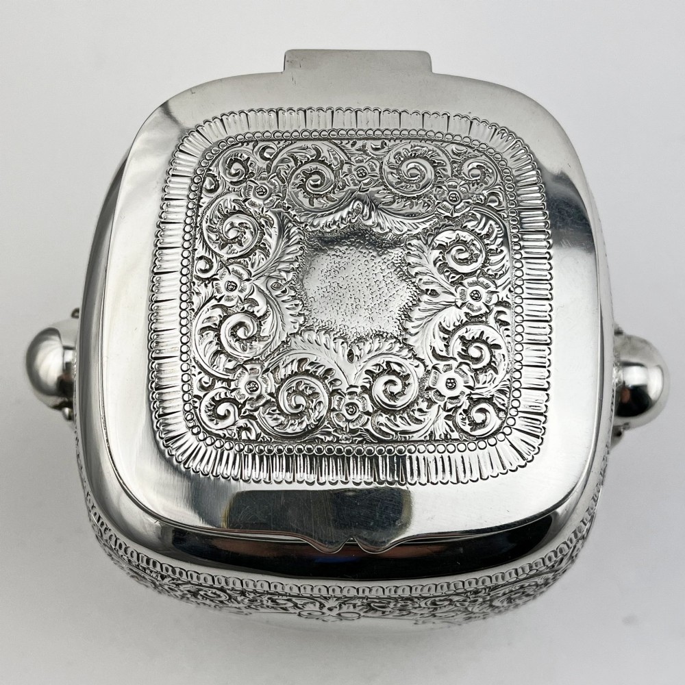 A silver-plated rectangular tea caddy featuring elaborate floral engraving, side handles, and a central blank cartouche. View II