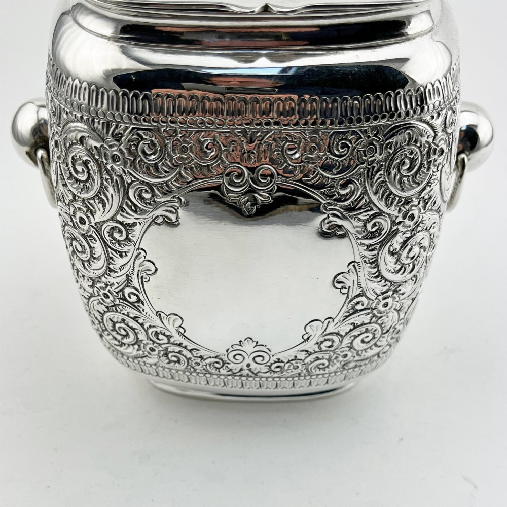 A silver-plated rectangular tea caddy featuring elaborate floral engraving, side handles, and a central blank cartouche. View III