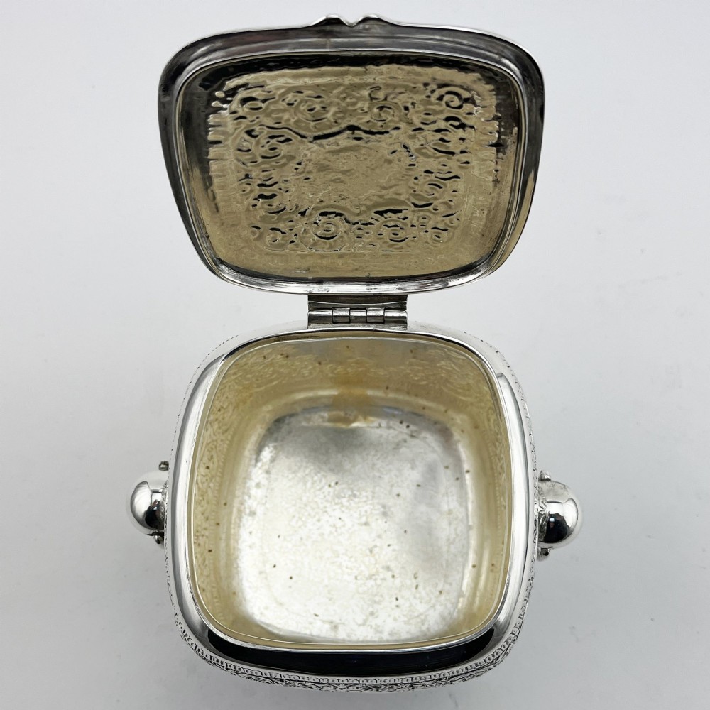 A silver-plated rectangular tea caddy featuring elaborate floral engraving, side handles, and a central blank cartouche. View V