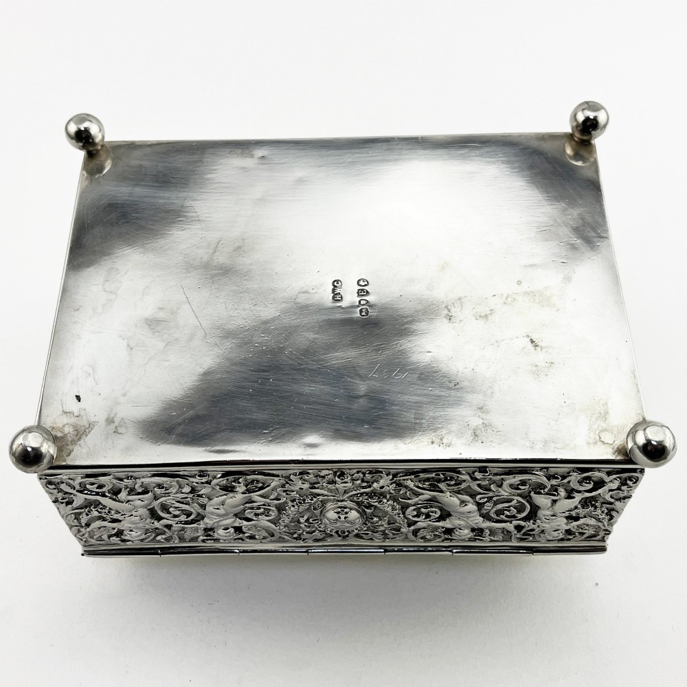 A rectangular Victorian sterling silver trinket box decorated with detailed repousse cherubs and floral scrolls, supported by four ball feet. View IX