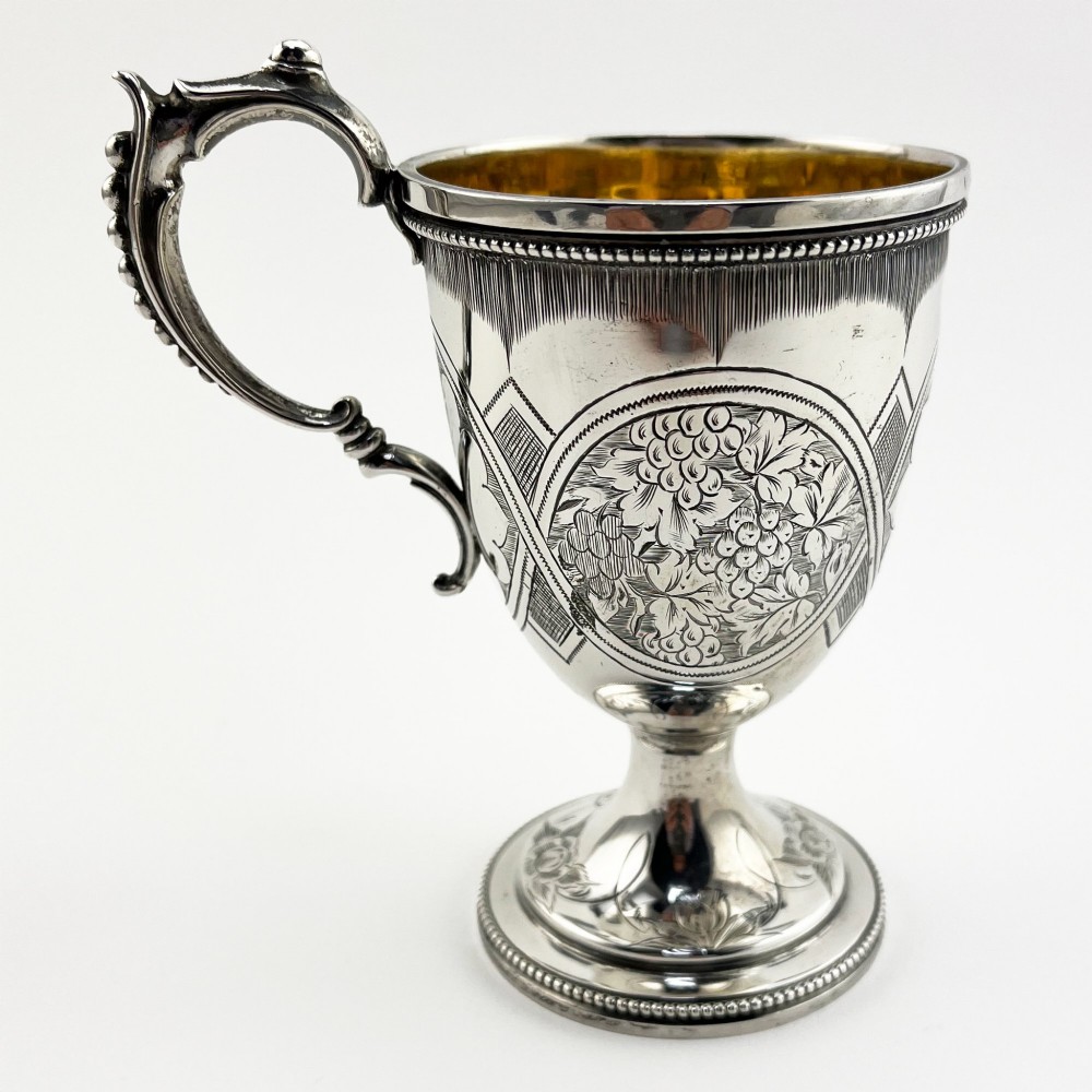 An antique Victorian sterling silver christening mug featuring a pedestal base, an ornate scroll handle, hand-engraved grapevine motifs, and a gilt interior lining.
