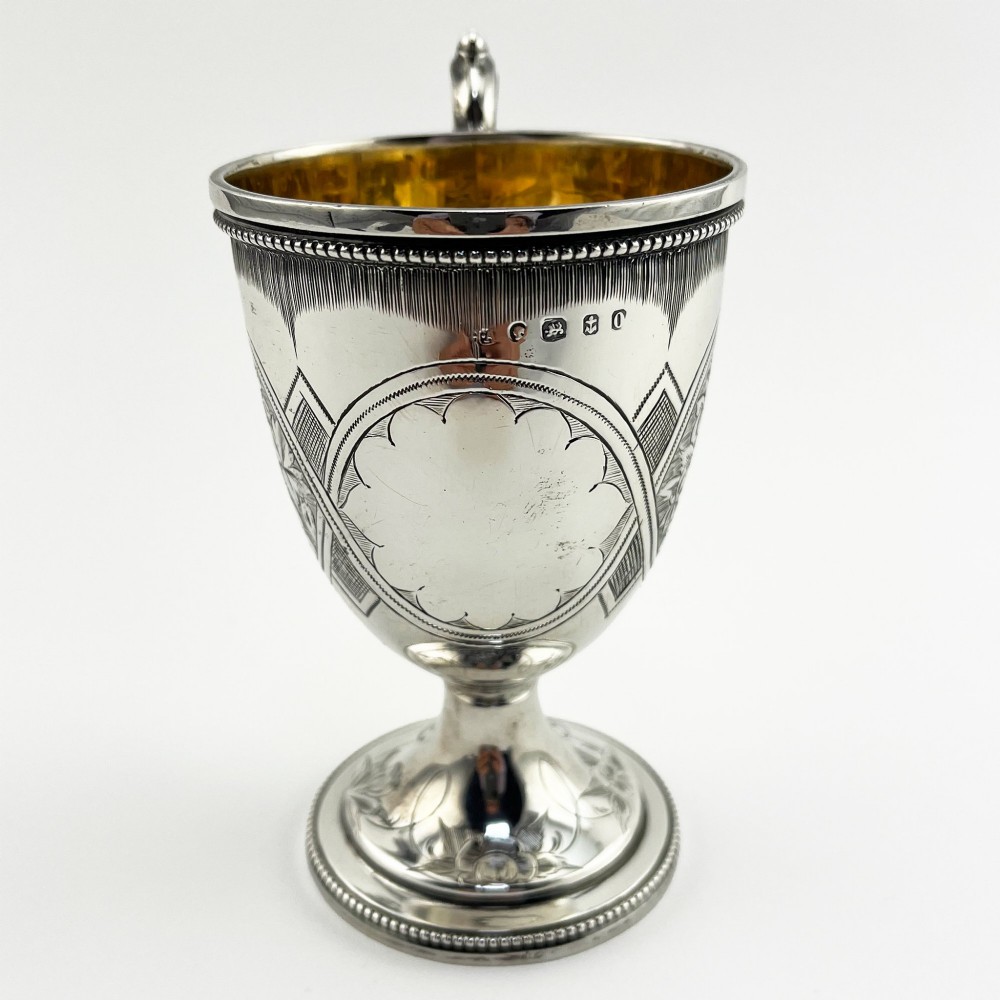 An antique Victorian sterling silver christening mug featuring a pedestal base, an ornate scroll handle, hand-engraved grapevine motifs, and a gilt interior lining. View II