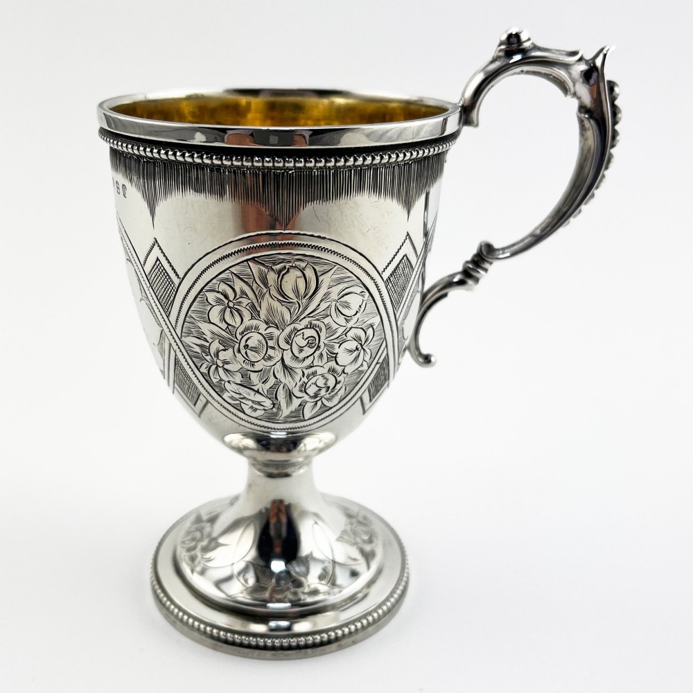 An antique Victorian sterling silver christening mug featuring a pedestal base, an ornate scroll handle, hand-engraved grapevine motifs, and a gilt interior lining. View III