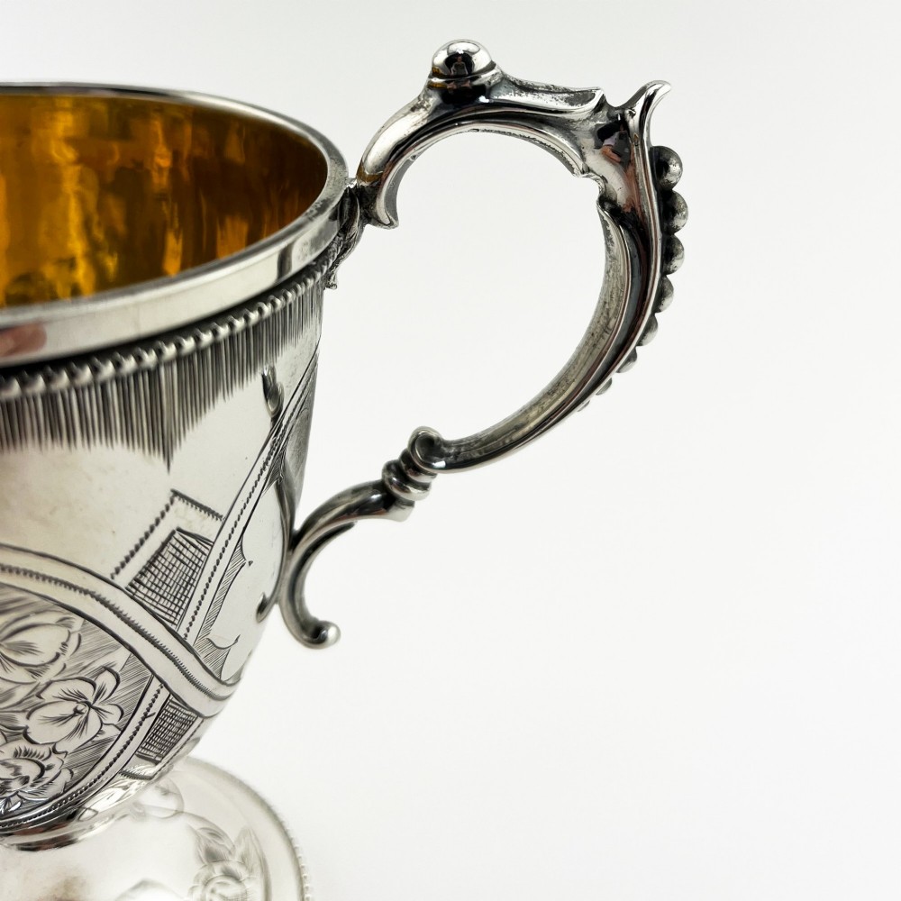 An antique Victorian sterling silver christening mug featuring a pedestal base, an ornate scroll handle, hand-engraved grapevine motifs, and a gilt interior lining. View IV