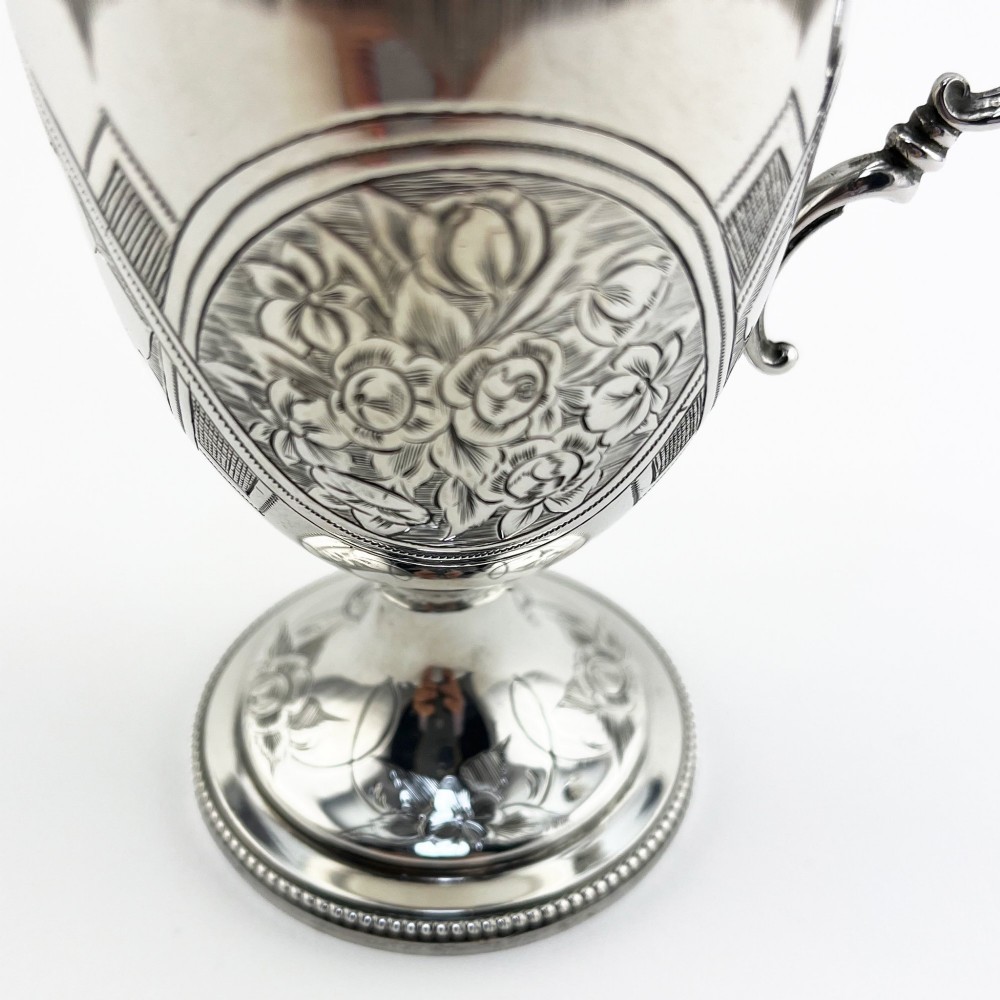 An antique Victorian sterling silver christening mug featuring a pedestal base, an ornate scroll handle, hand-engraved grapevine motifs, and a gilt interior lining. View V
