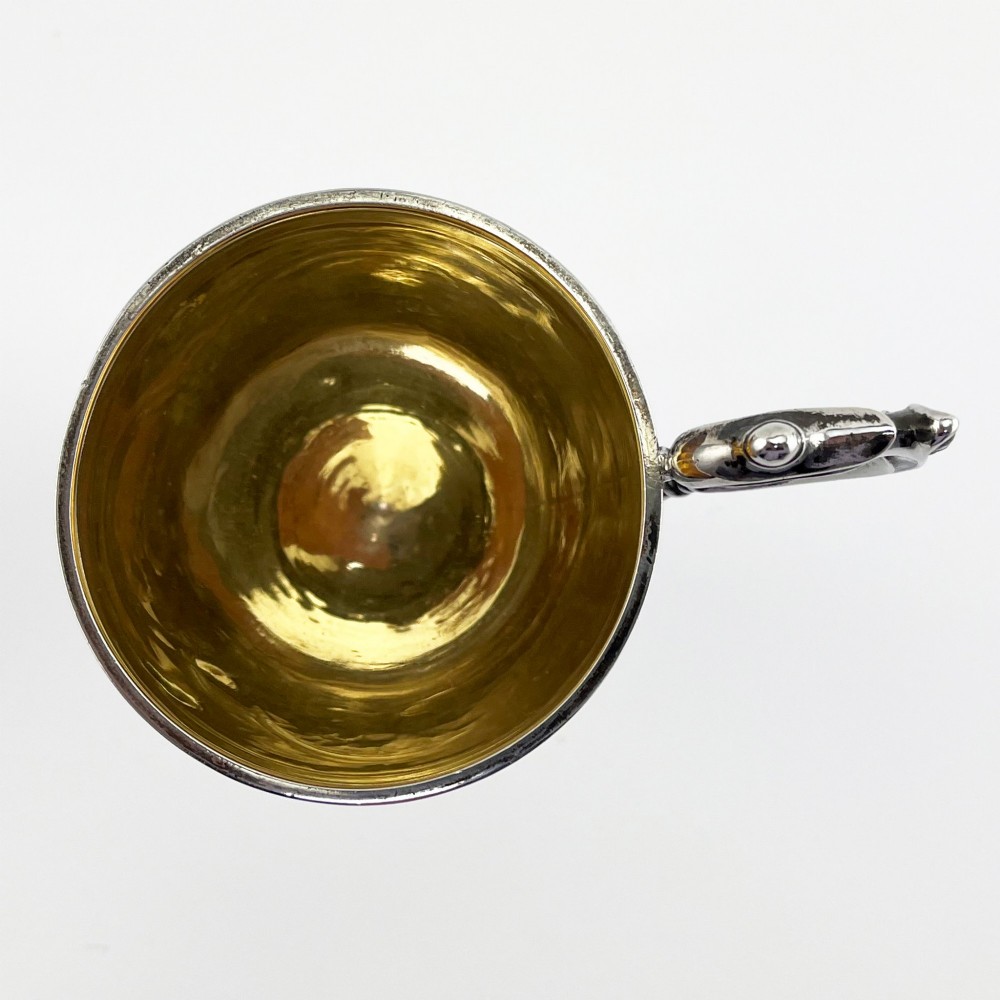 An antique Victorian sterling silver christening mug featuring a pedestal base, an ornate scroll handle, hand-engraved grapevine motifs, and a gilt interior lining. View VI