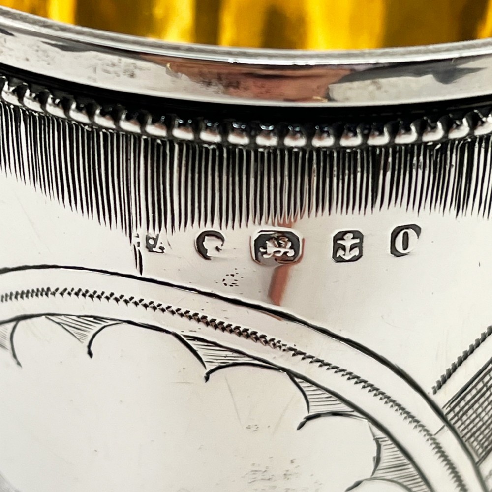 An antique Victorian sterling silver christening mug featuring a pedestal base, an ornate scroll handle, hand-engraved grapevine motifs, and a gilt interior lining. View VII