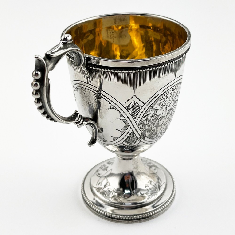 An antique Victorian sterling silver christening mug featuring a pedestal base, an ornate scroll handle, hand-engraved grapevine motifs, and a gilt interior lining. View VIII