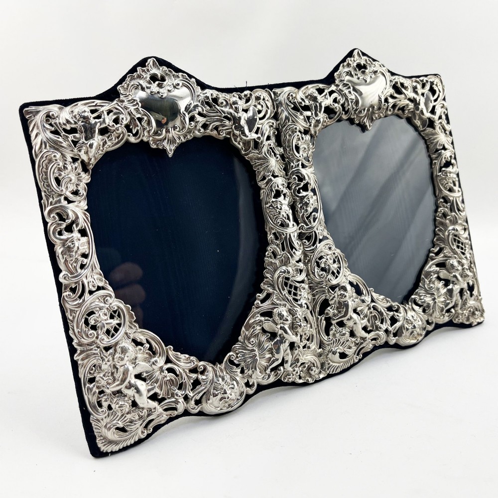 An ornate Victorian sterling silver double photo frame featuring elaborate pierced scrollwork, cherubs, and two heart-shaped apertures backed with dark velvet.