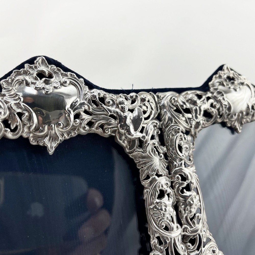 An ornate Victorian sterling silver double photo frame featuring elaborate pierced scrollwork, cherubs, and two heart-shaped apertures backed with dark velvet. View II