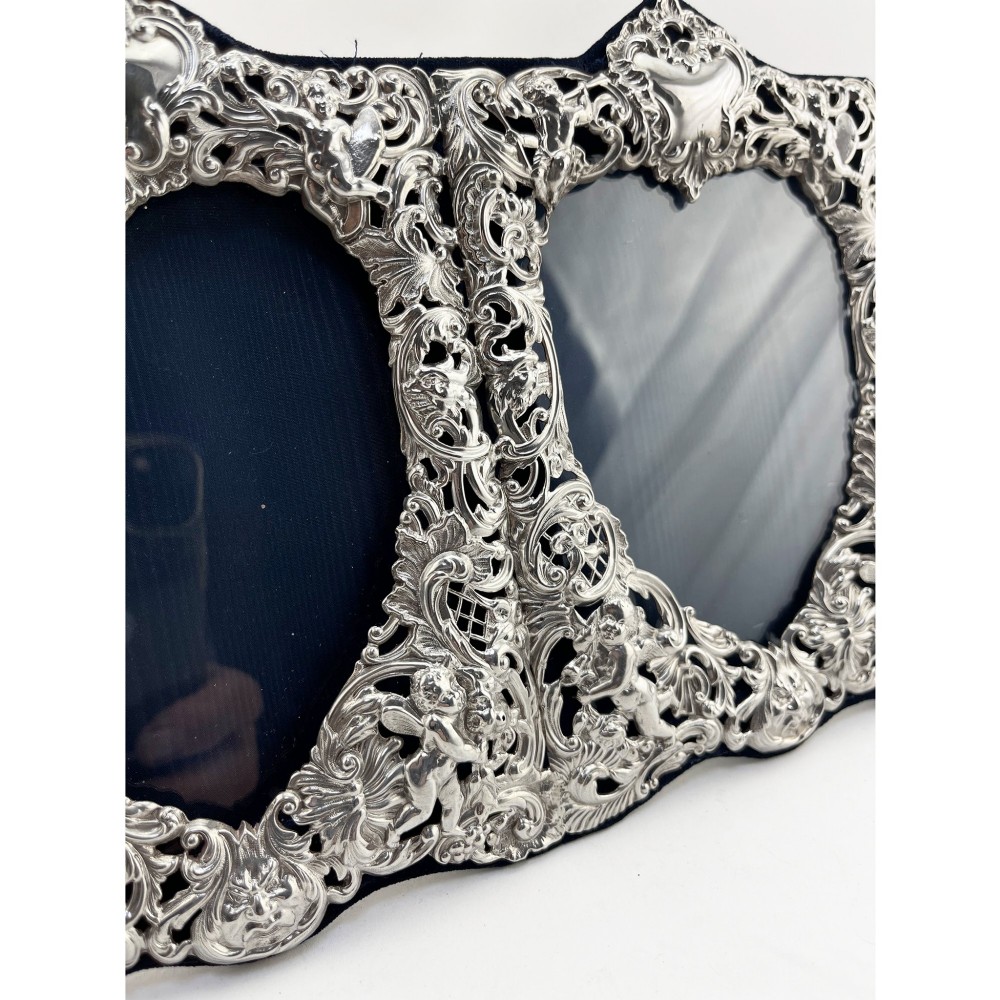 An ornate Victorian sterling silver double photo frame featuring elaborate pierced scrollwork, cherubs, and two heart-shaped apertures backed with dark velvet. View V