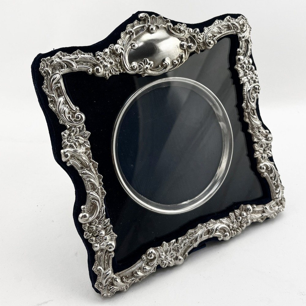 An ornate Edwardian sterling silver picture frame featuring chased floral scrollwork, a blank top cartouche, and a dark velvet mount with a central circular aperture.
