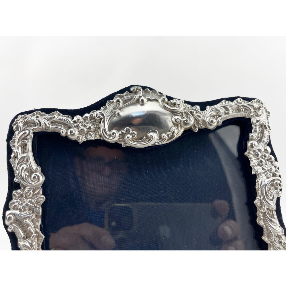An ornate Edwardian sterling silver picture frame featuring chased floral scrollwork, a blank top cartouche, and a dark velvet mount with a central circular aperture. View V