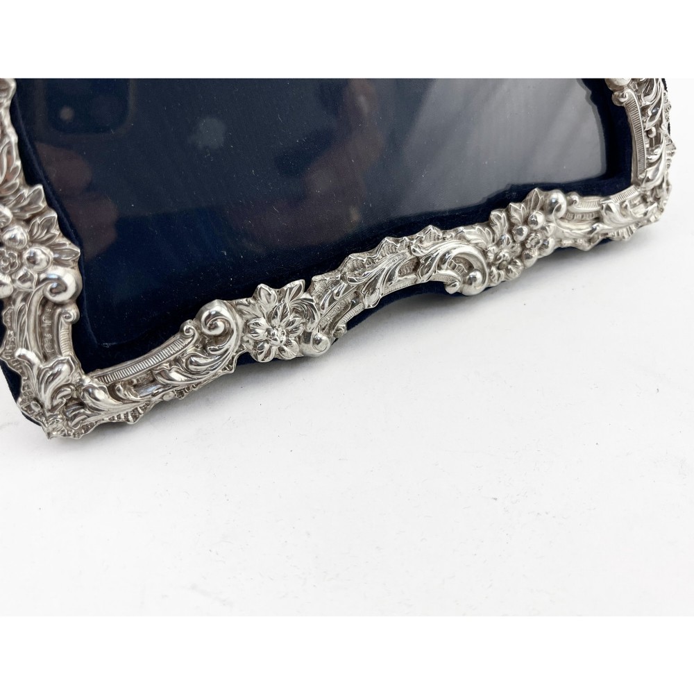 An ornate Edwardian sterling silver picture frame featuring chased floral scrollwork, a blank top cartouche, and a dark velvet mount with a central circular aperture. View VI