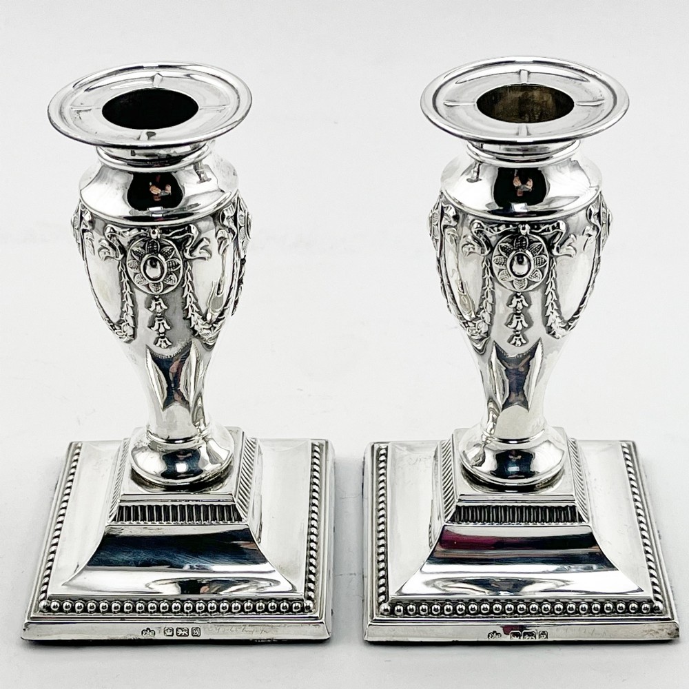 A pair of hallmarked Edwardian sterling silver candlesticks featuring urn-shaped stems with swag motifs and stepped beaded square bases.