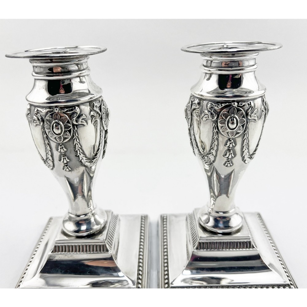 A pair of hallmarked Edwardian sterling silver candlesticks featuring urn-shaped stems with swag motifs and stepped beaded square bases. View V