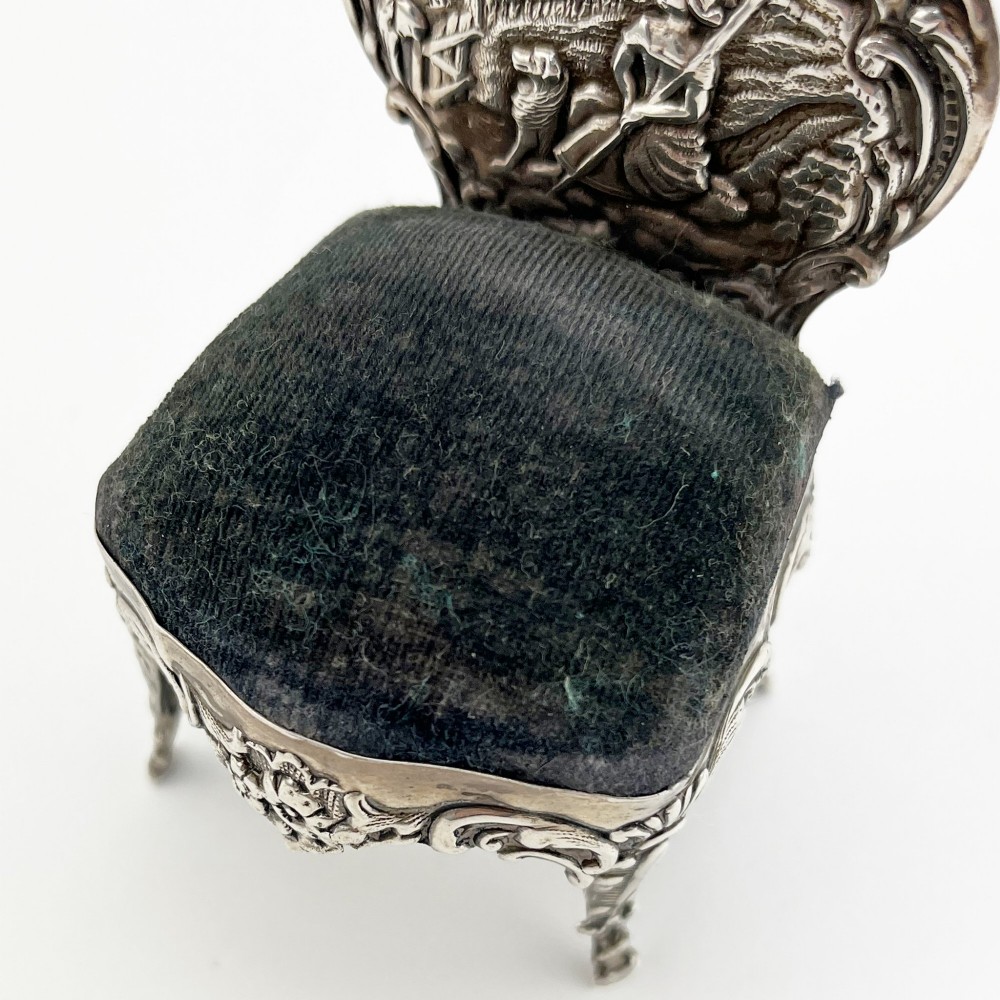 A miniature sterling silver pin cushion crafted in the shape of an ornate chair with an embossed backrest and dark velvet seat pad. View III