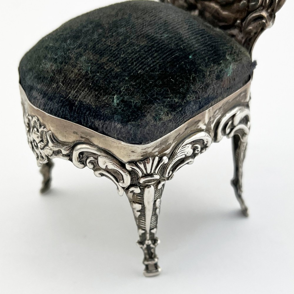 A miniature sterling silver pin cushion crafted in the shape of an ornate chair with an embossed backrest and dark velvet seat pad. View IV