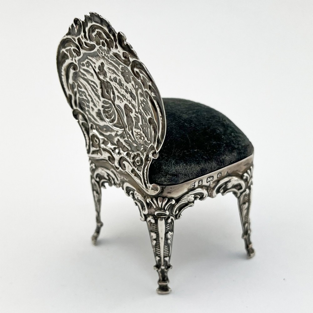 A miniature sterling silver pin cushion crafted in the shape of an ornate chair with an embossed backrest and dark velvet seat pad. View V
