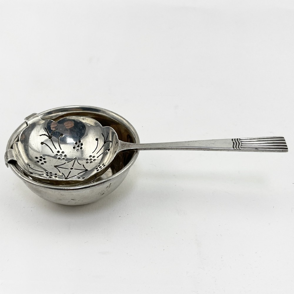 A sterling silver tea strainer featuring star perforations resting inside a circular silver drip bowl.