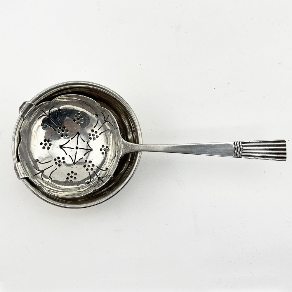 A sterling silver tea strainer featuring star perforations resting inside a circular silver drip bowl. View II