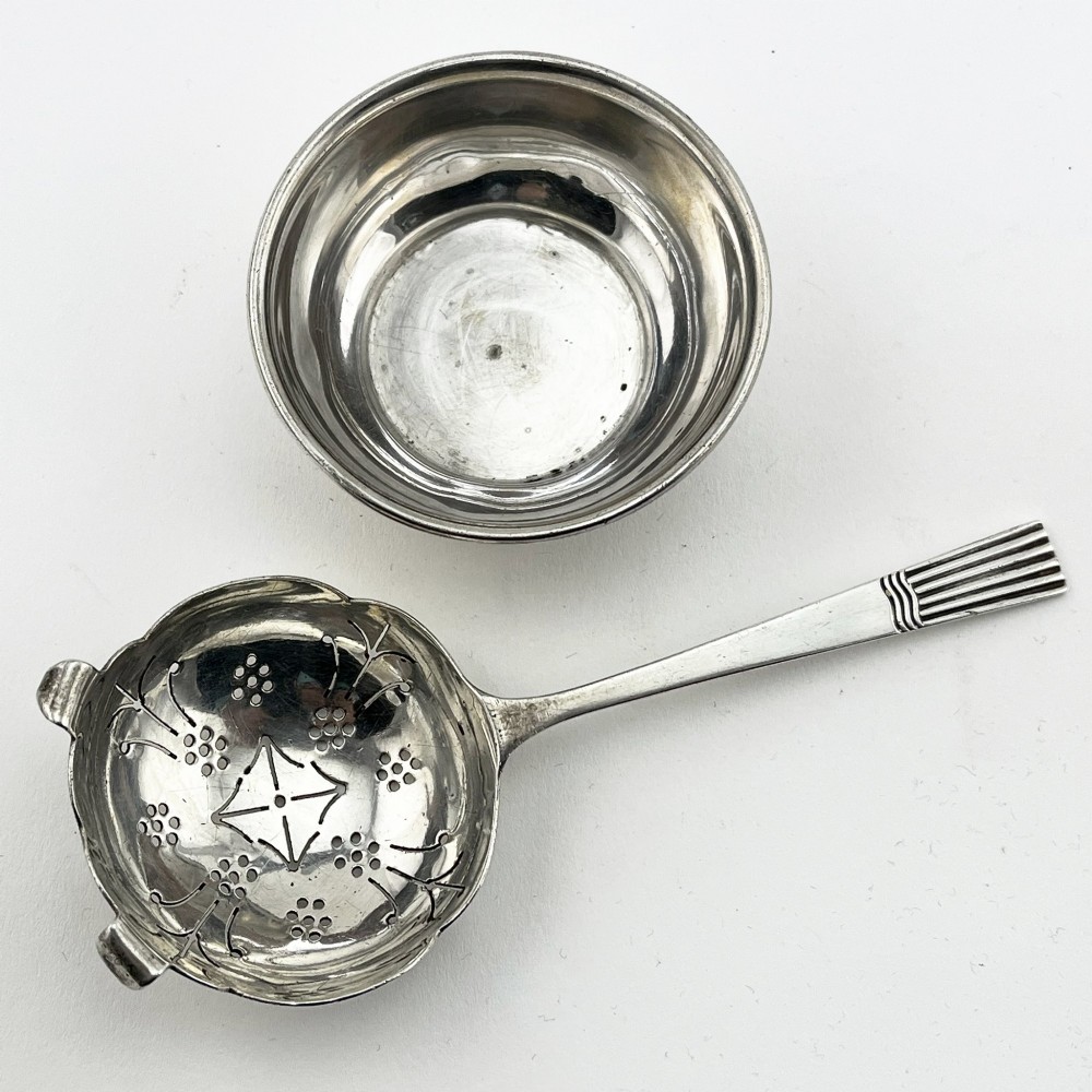 A sterling silver tea strainer featuring star perforations resting inside a circular silver drip bowl. View III