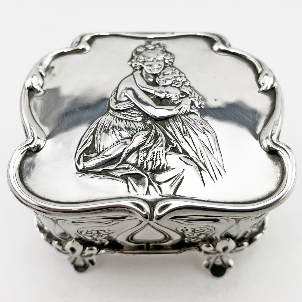 An Edwardian sterling silver trinket box featuring a raised relief depiction of a mother embracing a child on the hinged lid, set on scrolled silver feet.