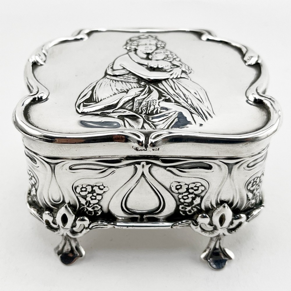 An Edwardian sterling silver trinket box featuring a raised relief depiction of a mother embracing a child on the hinged lid, set on scrolled silver feet. View II
