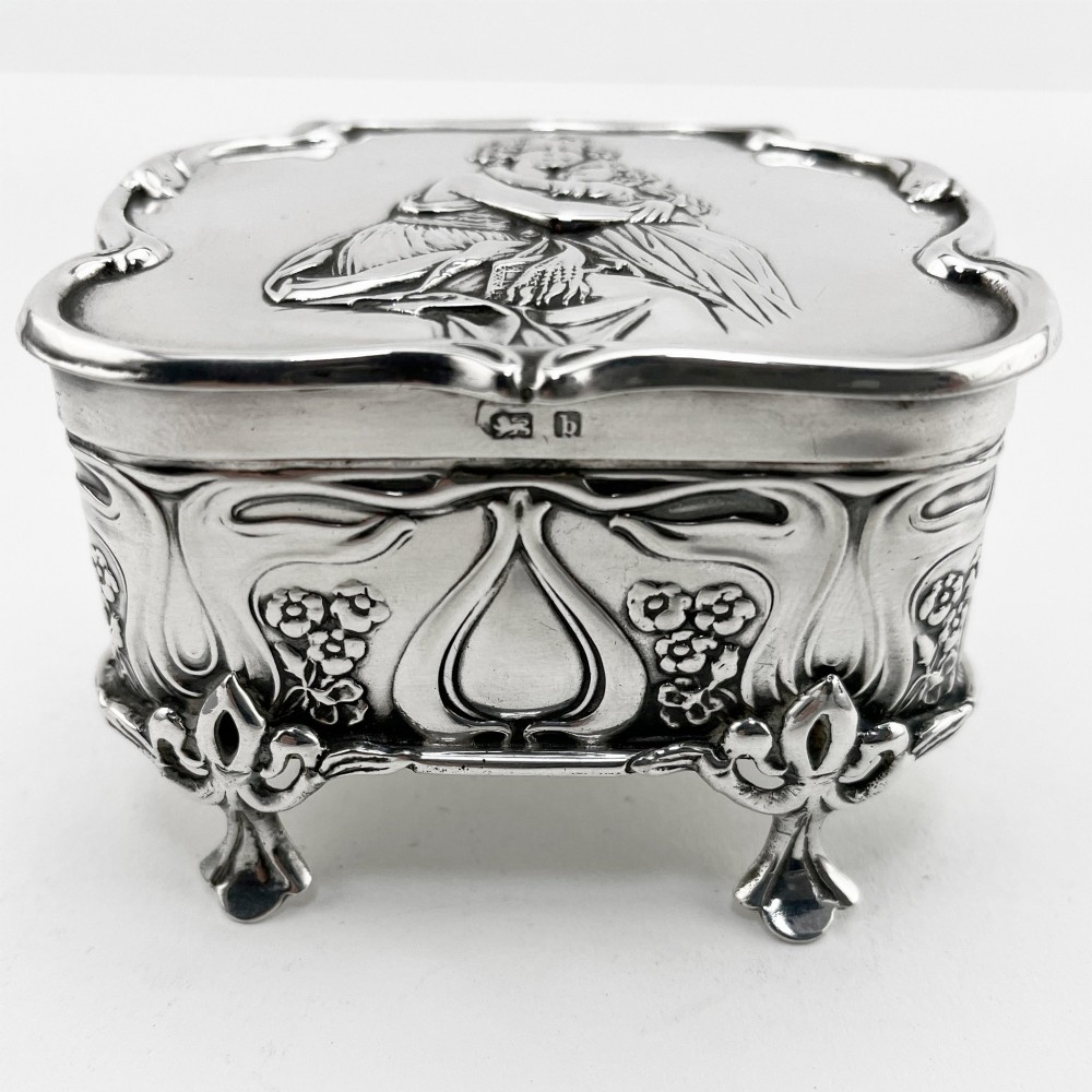 An Edwardian sterling silver trinket box featuring a raised relief depiction of a mother embracing a child on the hinged lid, set on scrolled silver feet. View III