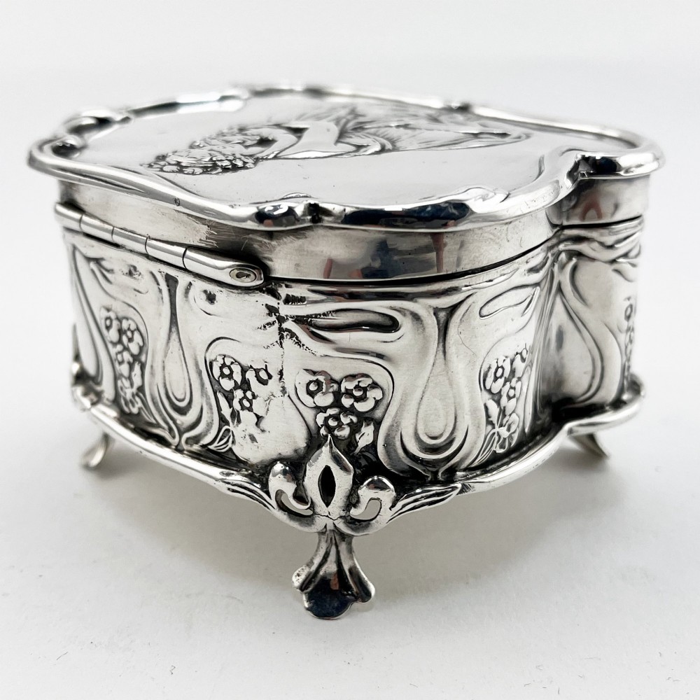 An Edwardian sterling silver trinket box featuring a raised relief depiction of a mother embracing a child on the hinged lid, set on scrolled silver feet. View V