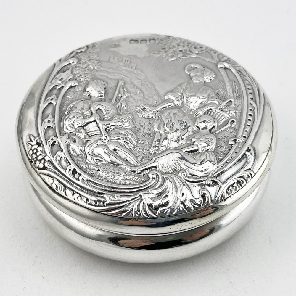 A circular sterling silver trinket box featuring a repousse relief on the lid depicting a seated bagpipe player and figures in a landscape.