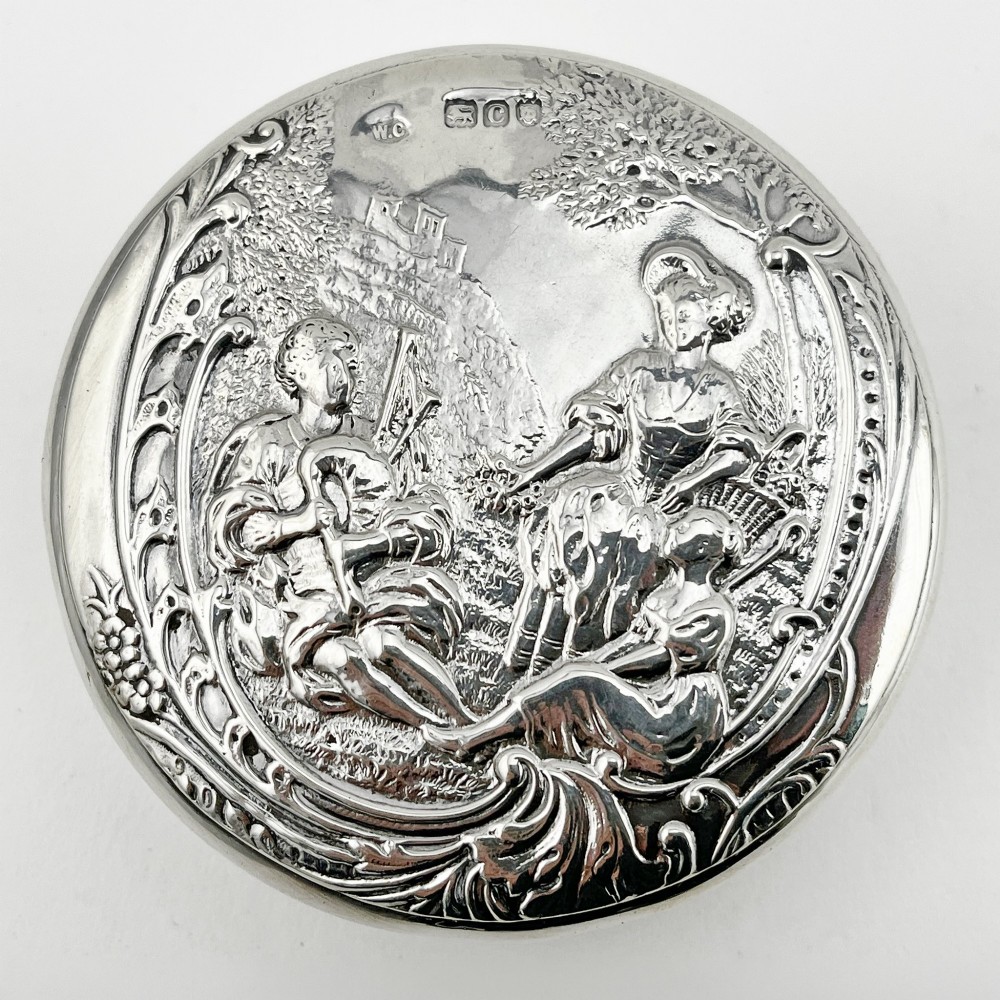 A circular sterling silver trinket box featuring a repousse relief on the lid depicting a seated bagpipe player and figures in a landscape. View II