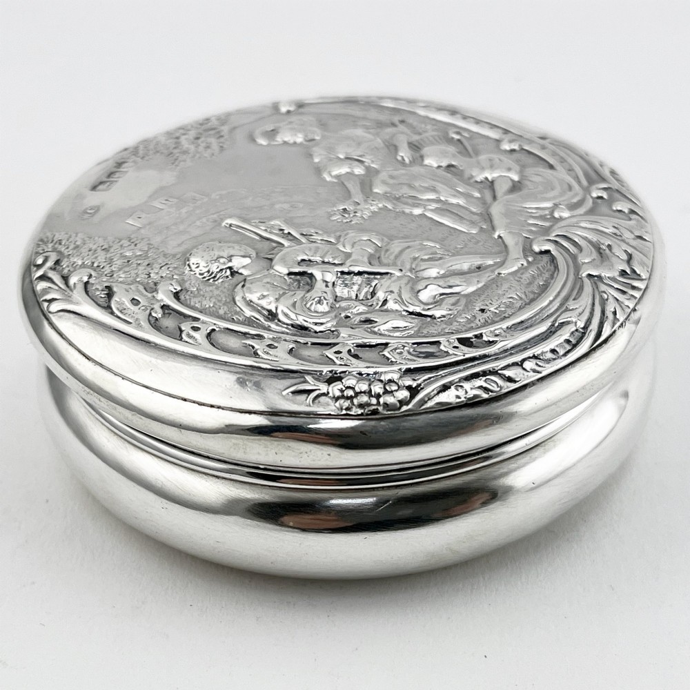 A circular sterling silver trinket box featuring a repousse relief on the lid depicting a seated bagpipe player and figures in a landscape. View III