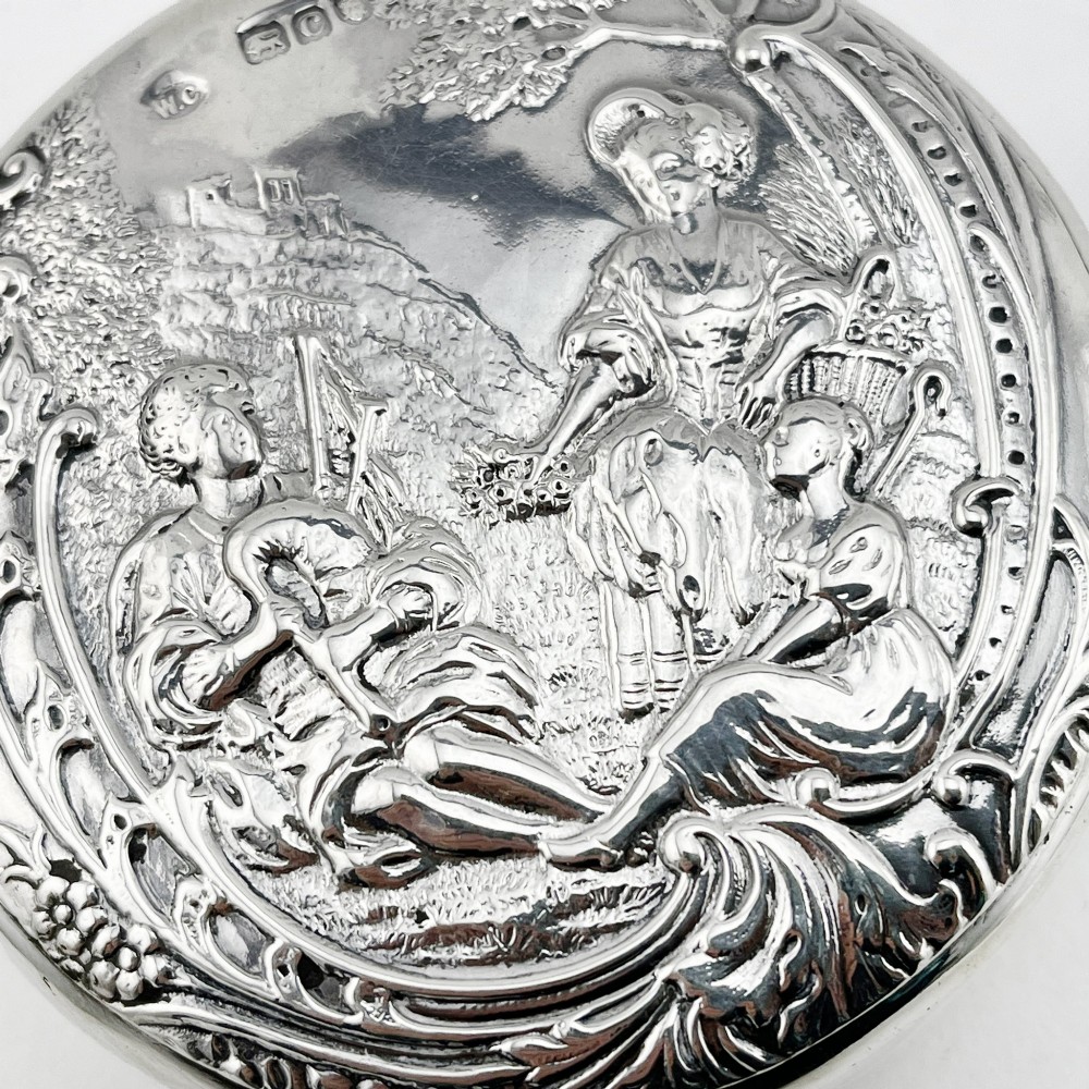 A circular sterling silver trinket box featuring a repousse relief on the lid depicting a seated bagpipe player and figures in a landscape. View IV