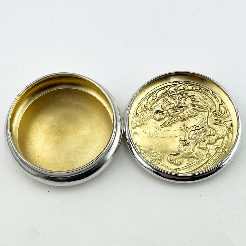 A circular sterling silver trinket box featuring a repousse relief on the lid depicting a seated bagpipe player and figures in a landscape. View V