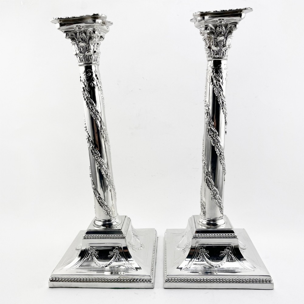 A pair of silver-plated column candlesticks featuring Corinthian capitals, spiral foliate garlands along the shafts, and square stepped bases.