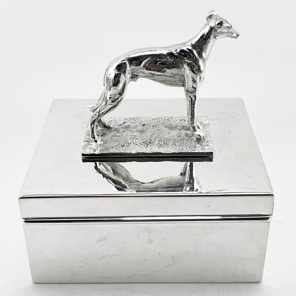 A rectangular silver-plated box featuring a polished finish and a sculpted figure of a standing greyhound attached to the lid. View II