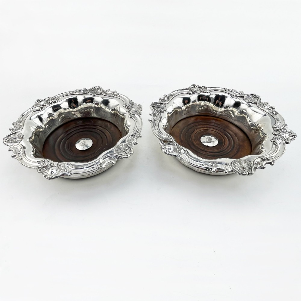A pair of circular silver plated wine coasters featuring shell and scroll borders with turned dark wood bases and silver central bosses.