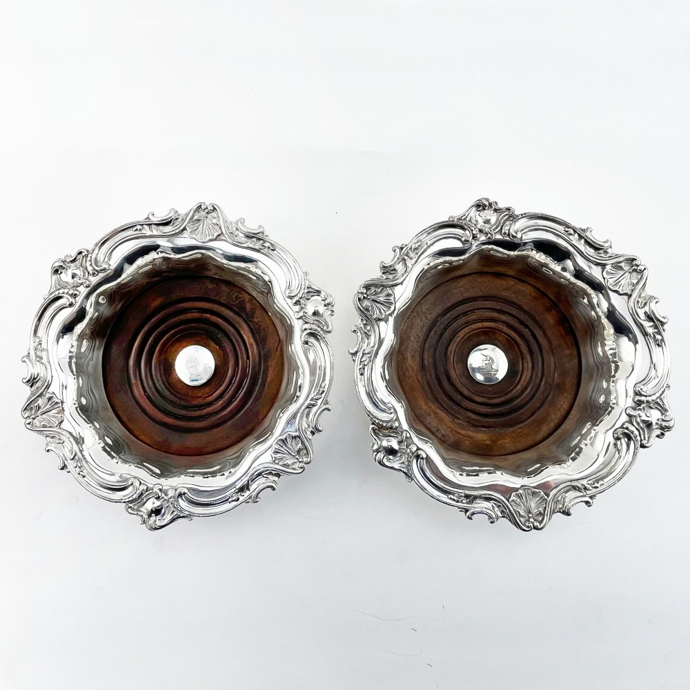 A pair of circular silver plated wine coasters featuring shell and scroll borders with turned dark wood bases and silver central bosses. View II