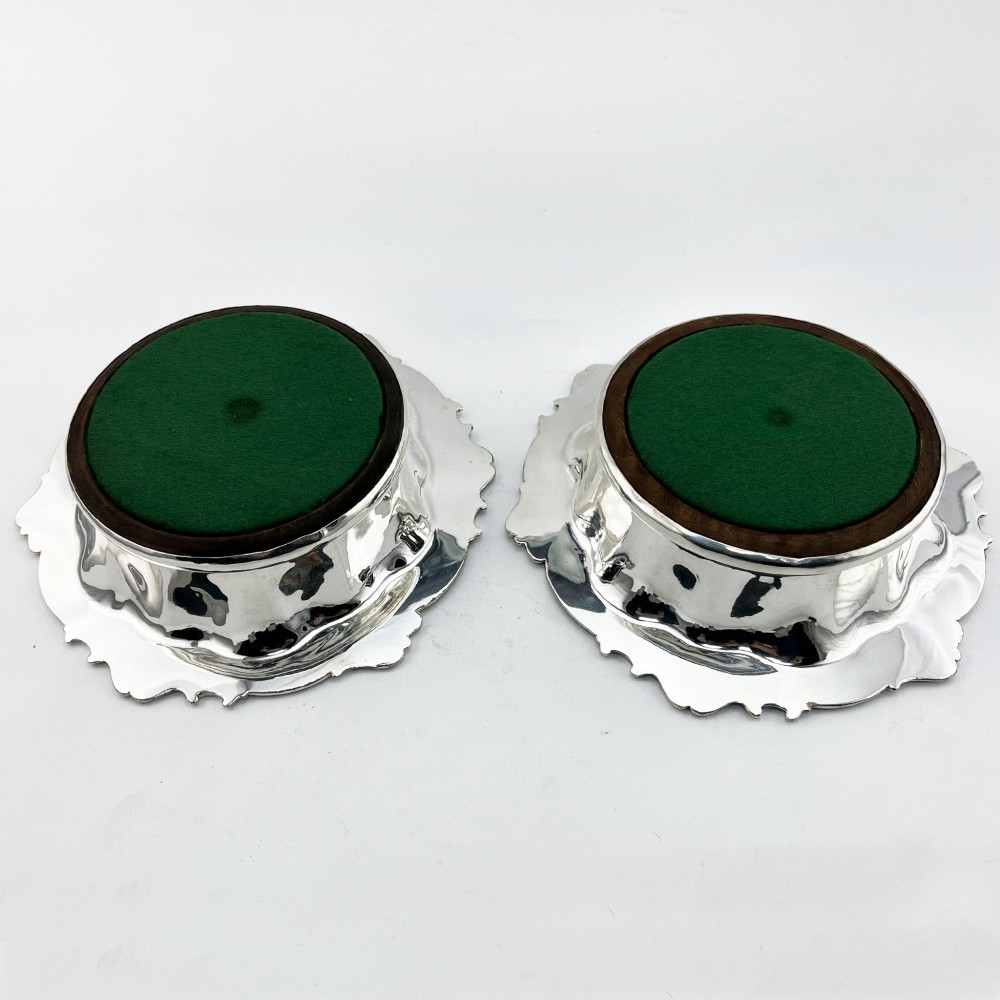 A pair of circular silver plated wine coasters featuring shell and scroll borders with turned dark wood bases and silver central bosses. View VI